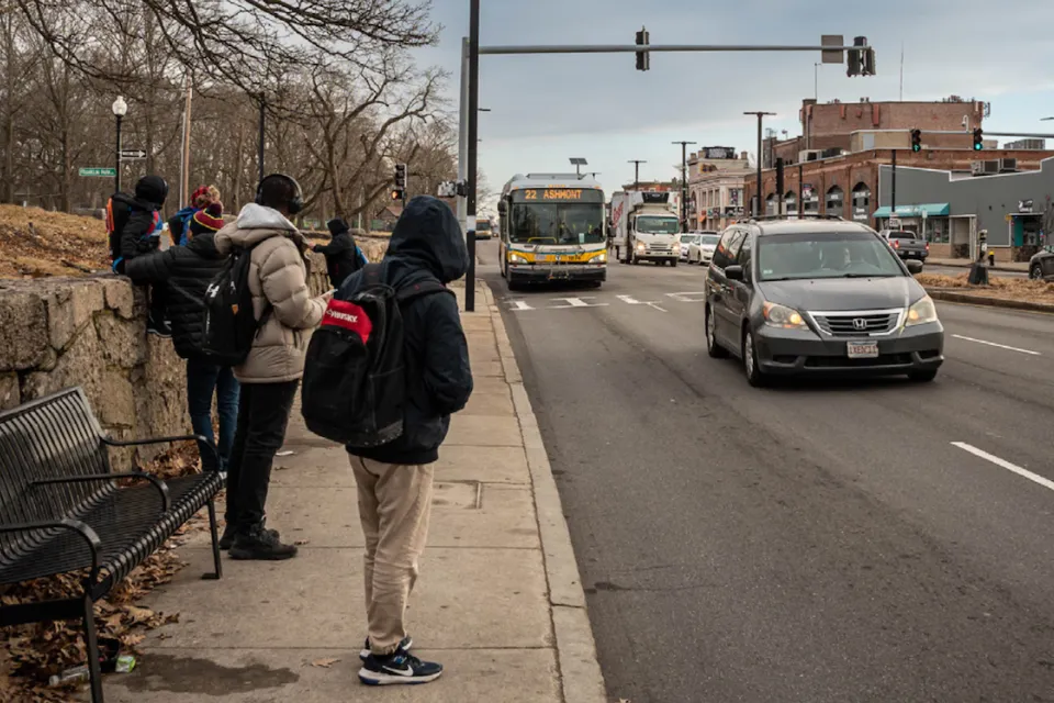 People in hoodies and jackets waiting for oncoming bus at sidewalk bus stop in Boston, Massachusetts.